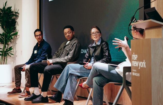 A panel of four diverse professionals engaged in a discussion at Rise New York, hosted by Barclays. The panelists are seated on stage, holding microphones, while the moderator, seated at a podium, gestures as they speak. The background features a large screen displaying event branding, and the setting has a modern, professional atmosphere.