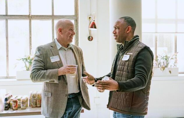 Two men having a friendly conversation at a networking event. One wears a light blazer and holds a drink, while the other wears a dark quilted vest and gestures while speaking. A table with drinks is visible in the background by large windows.