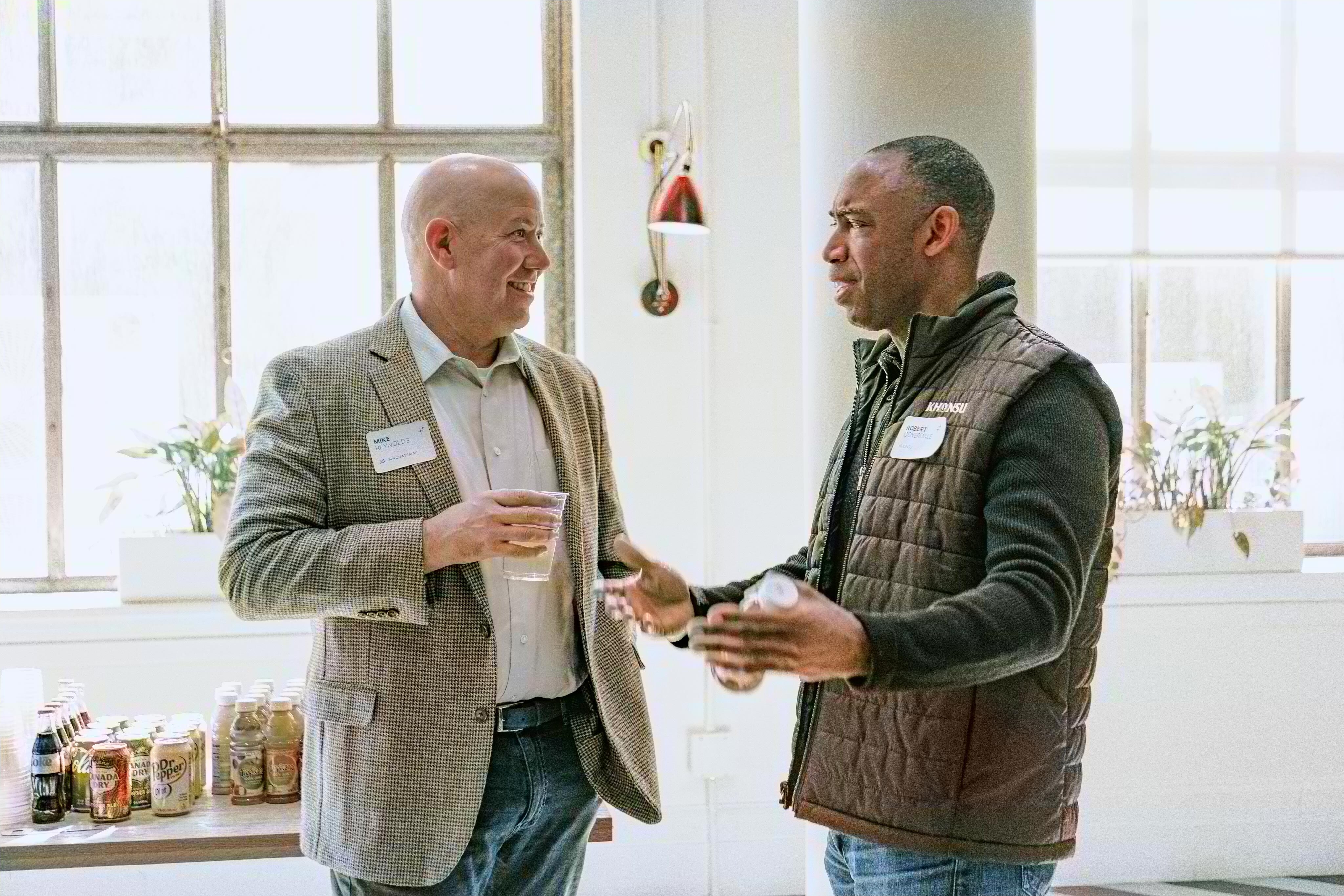 Two men having a friendly conversation at a networking event. One wears a light blazer and holds a drink, while the other wears a dark quilted vest and gestures while speaking. A table with drinks is visible in the background by large windows.